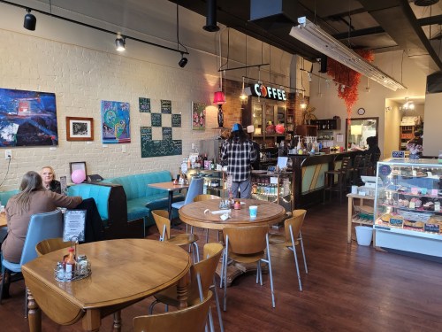 A cozy café interior with wooden tables, colorful artwork, and a coffee counter. Customers enjoy drinks and conversation.