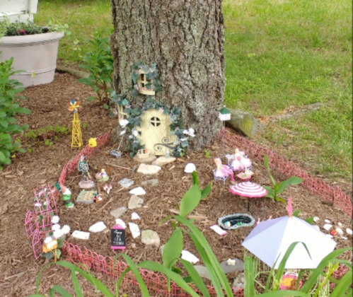 A whimsical fairy garden at the base of a tree, featuring a tiny house, decorations, and colorful figurines.