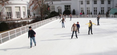 A group of people ice skating on an outdoor rink surrounded by white buildings and trees.