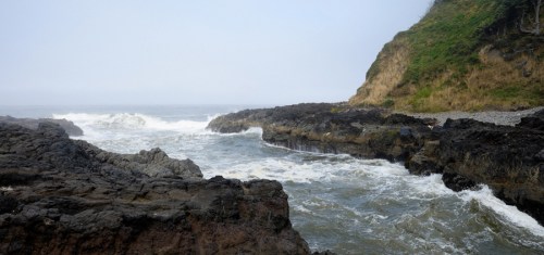 A rocky coastline with waves crashing against the shore, surrounded by lush green hills under a foggy sky.