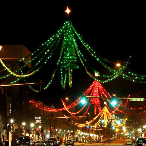 Colorful holiday lights in the shape of a Christmas tree adorn a city street at night, creating a festive atmosphere.