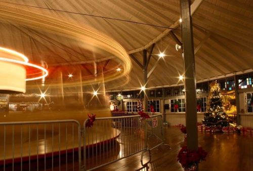 A festive carousel spins inside a warmly lit pavilion, decorated with holiday lights and a Christmas tree.
