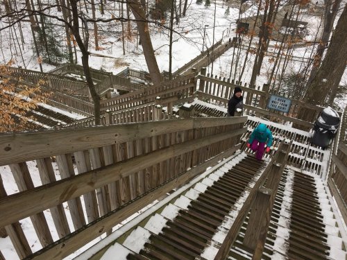 A snowy wooden staircase winds through a forest, with two people climbing up and a sign visible in the background.