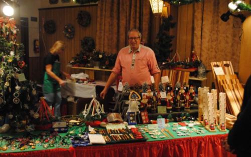 A vendor stands at a festive market table filled with handmade crafts and decorations, with a Christmas tree in the background.