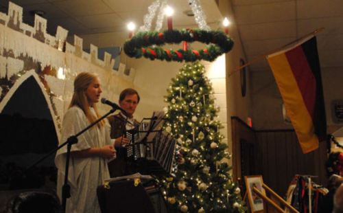 A young girl in a white dress sings near a decorated Christmas tree, with a man playing an accordion beside her.