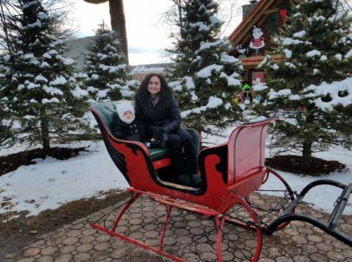 A woman and a child sit in a red sleigh surrounded by snow-covered trees and a festive cabin in the background.