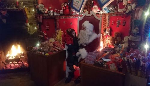A child sits on Santa's lap in a festive room filled with toys, decorations, and a cozy fireplace.