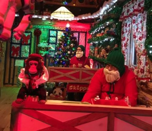 Three festive figures, including a monkey and two Santa Claus figures, are seated in a colorful, decorated holiday setting.