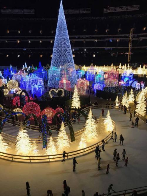 A festive winter scene with colorful lights, a large tree, and people skating on ice surrounded by illuminated decorations.