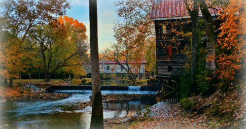 A rustic wooden mill beside a river, surrounded by vibrant autumn foliage and a gentle waterfall.