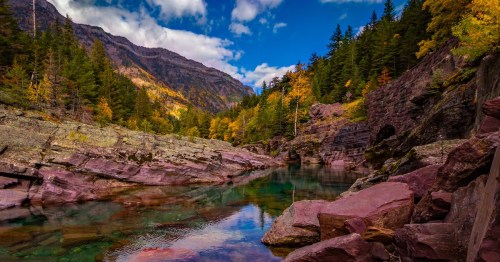 A serene landscape featuring a clear river surrounded by colorful autumn trees and rocky mountains under a blue sky.