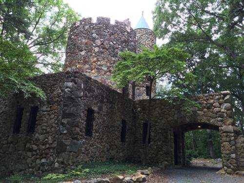 A stone castle with a turret, surrounded by trees and a gravel path leading to an archway entrance.