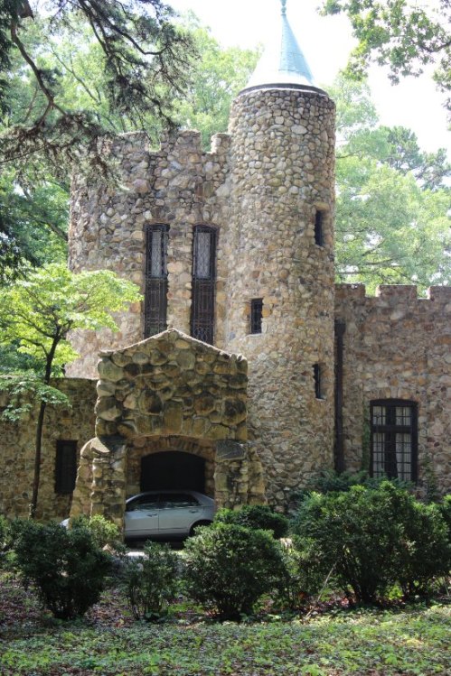 A stone castle with a turret, surrounded by trees and greenery, featuring an arched entrance and a parked car.