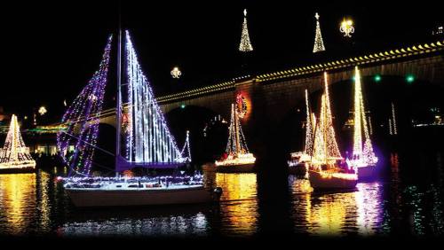 Colorful, illuminated boats decorated for the holidays float on a dark river, with festive lights reflecting on the water.