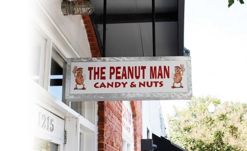 Sign for "The Peanut Man" featuring cartoon peanuts, advertising candy and nuts, hanging above a storefront.