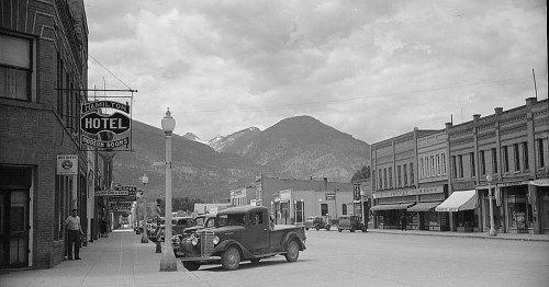 Black and white photo of a street scene with vintage cars, a hotel sign, and mountains in the background.