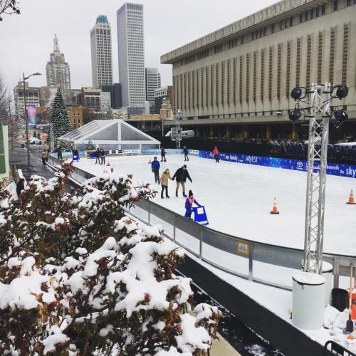 A winter scene featuring an ice skating rink surrounded by snow, with city buildings in the background.
