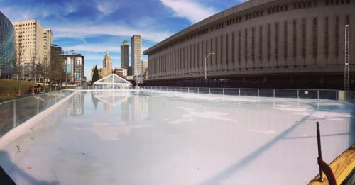 A large outdoor ice skating rink surrounded by city buildings under a clear blue sky.