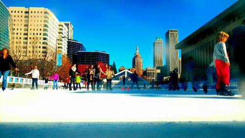 A bustling ice skating rink in a city, with people skating and tall buildings in the background under a clear blue sky.