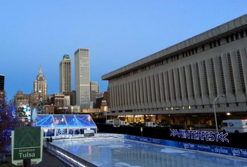 A winter ice skating rink in downtown Tulsa, with city skyscrapers and festive lights in the background.