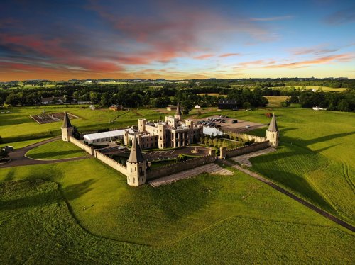 Aerial view of a castle surrounded by green fields and a colorful sunset sky.