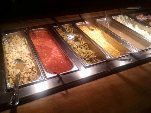 A buffet with five trays of various pasta dishes, including sauces and sides, under warm lighting.