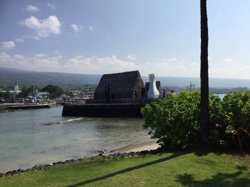 A scenic view of a coastal area featuring a thatched-roof building, palm trees, and calm waters under a blue sky.