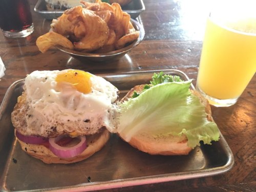 A burger with a fried egg, lettuce, and onion on a bun, served with crispy potato chips and a glass of yellow drink.