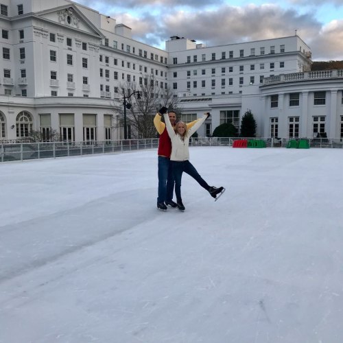 A couple ice skating on a rink, smiling and posing with their arms raised, surrounded by a scenic winter backdrop.