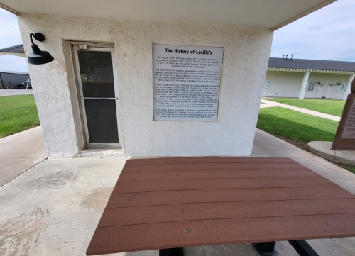 A building with a sign titled "The History of Lucille's" and a picnic table in front on a grassy area.