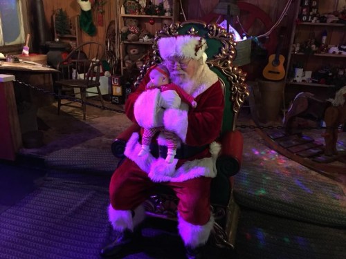 Santa Claus sits in a decorated chair, gently holding a baby in festive attire, surrounded by holiday decorations.