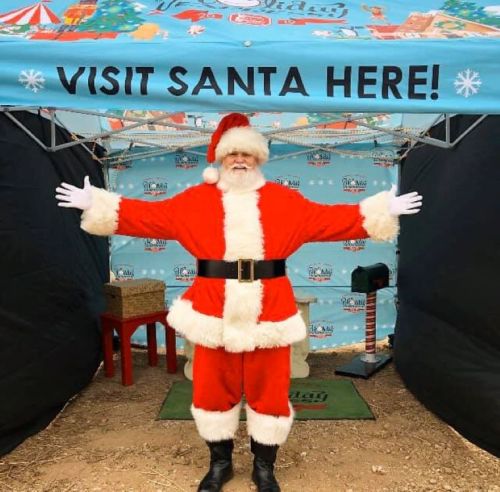 A cheerful Santa Claus stands with open arms in front of a festive "Visit Santa Here!" sign.