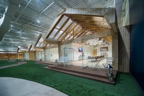 Modern indoor space with wooden architecture, glass walls, and green turf, featuring seating areas and a cozy atmosphere.