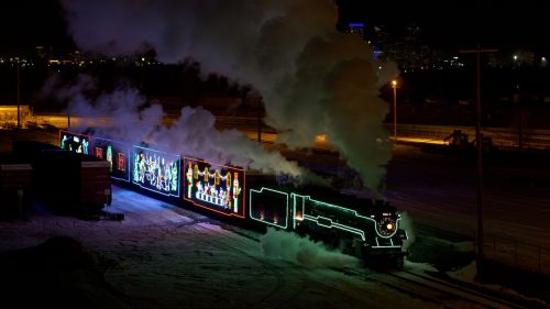 A brightly lit steam train with colorful decorations emits smoke at night, set against a city skyline.