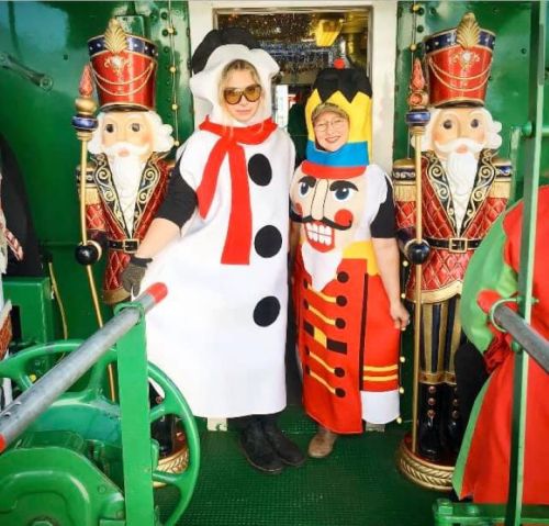 Two women in festive costumes stand between two nutcracker figures, smiling in a colorful holiday setting.