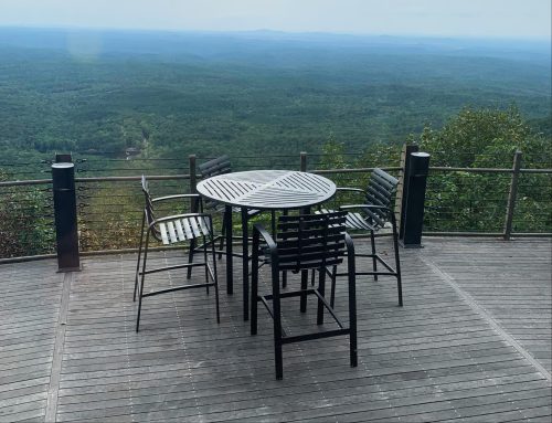 A round table with four chairs on a wooden deck overlooking a lush green mountain landscape.