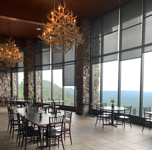 A spacious dining area with large windows, stone walls, and antler chandeliers, overlooking a scenic mountain view.
