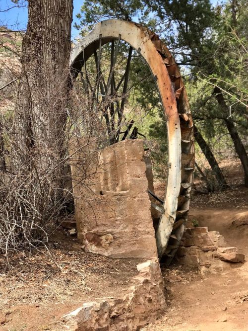 An old, rusted water wheel partially hidden by trees and brush, next to a stone structure in a natural setting.