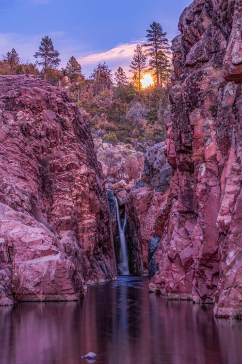 A serene canyon with pink rock formations, a waterfall, and a sunset peeking through the trees in the background.