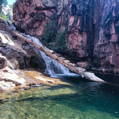 A serene waterfall cascades over rocks into a clear pool, surrounded by lush greenery and rugged cliffs.