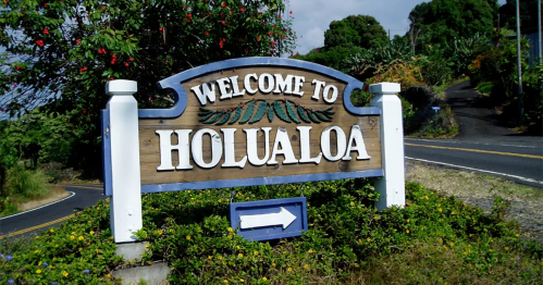 A wooden welcome sign for Holualoa, surrounded by greenery and flowers, with a road visible in the background.