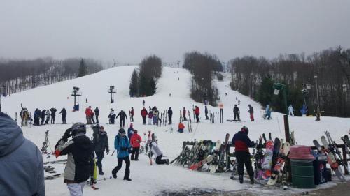 Skiers and snowboarders gather at a snowy slope, with equipment and ski lifts in the background on a cloudy day.