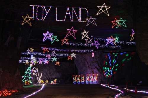 Colorful holiday lights spell "Toy Land" and feature stars, a dinosaur, and festive figures in a cave setting.