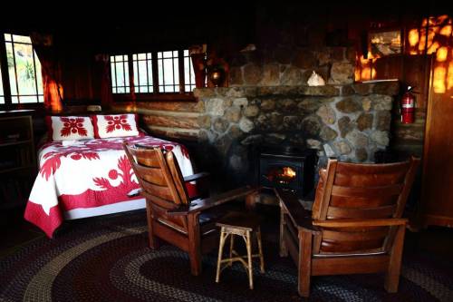 Cozy cabin interior with a stone fireplace, wooden chairs, and a bed adorned with a red and white quilt.