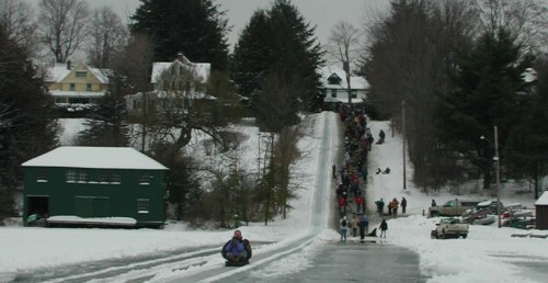 A snowy hill with people sledding down and walking up, surrounded by trees and houses in a winter landscape.