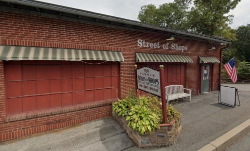 A brick building with a sign reading "Street of Shops," featuring green awnings and a bench in front. An American flag is visible.