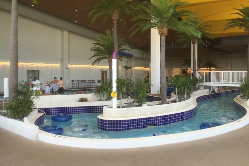 Indoor water park area with a lazy river, palm trees, and people enjoying the space. Bright and inviting atmosphere.