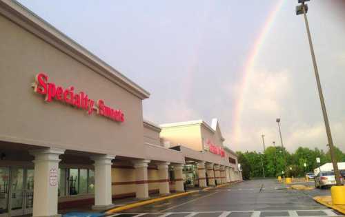 A storefront with "Specialty Snacks" signage under a rainbow after rain, with a mostly empty parking lot.