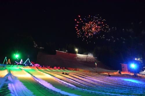 Colorful fireworks illuminate a snowy landscape at night, with vibrant lights on a sledding hill and festive decorations.