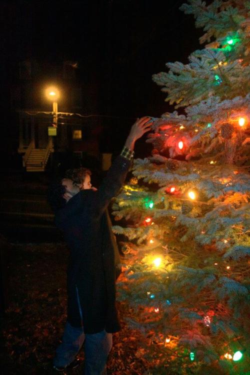 A person reaches up to decorate a brightly lit Christmas tree at night.